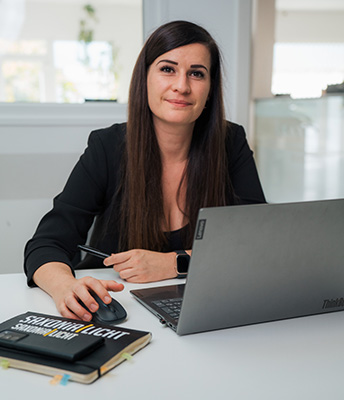 Eine Frau mit langen dunklen Haaren sitzt an einem Schreibtisch in einem Büro und benutzt einen Laptop. Sie hält eine Maus in der Hand und hat ein Notizbuch mit dem Titel „Schaulicht“ auf dem Schreibtisch liegen. Natürliches Licht strömt durch das Fenster und schafft einen inspirierenden Arbeitsplatz für ihre Rolle als Leitender Monteur.