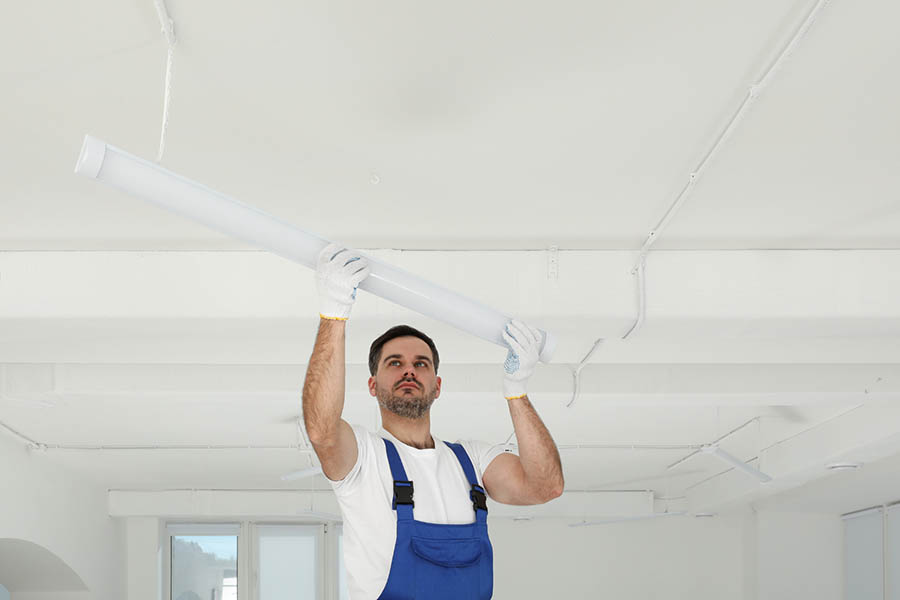 Ein Mann im blauen Overall installiert eine Leuchtstoffröhre an einer weißen Decke. Er benutzt beide Hände und trägt Handschuhe. Der Raum ist hell und wirkt modern und minimalistisch.