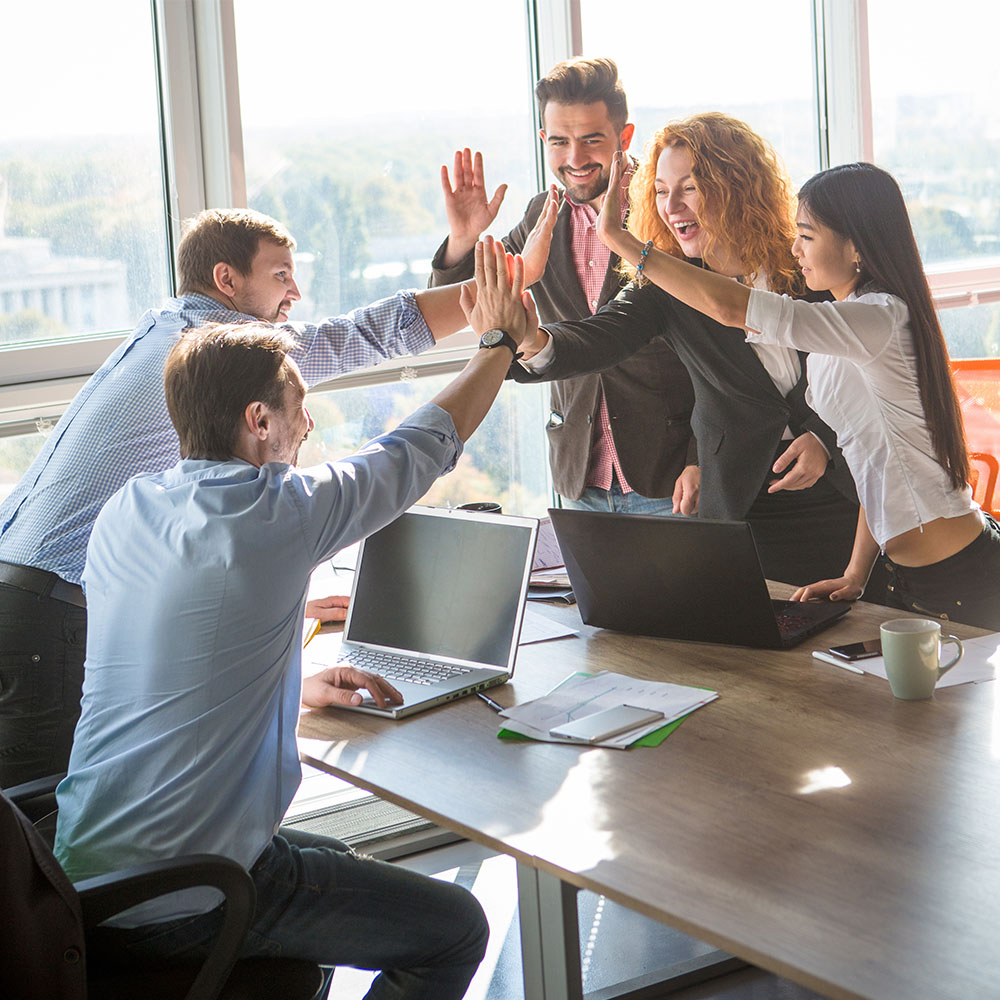 Eine Gruppe von fünf Personen in Business-Kleidung sitzt in einem hellen Büro um einen Tisch herum und gibt sich gegenseitig High Fives. Sie wirken enthusiastisch und fröhlich. Laptops, Papiere und eine Kaffeetasse liegen auf dem Tisch. Große Fenster lassen Tageslicht herein und zeigen ihren Erfolg in LED-Technik.