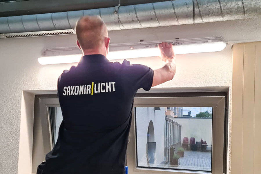 Eine Person, die ein „Saxonia Licht“-Shirt trägt, installiert oder justiert eine Leuchtstoffröhre an einer Wand über einem großen Fenster. Der Raum scheint sich in einem Gebäude zu befinden, mit sichtbaren Leitungen in der Nähe der Decke.